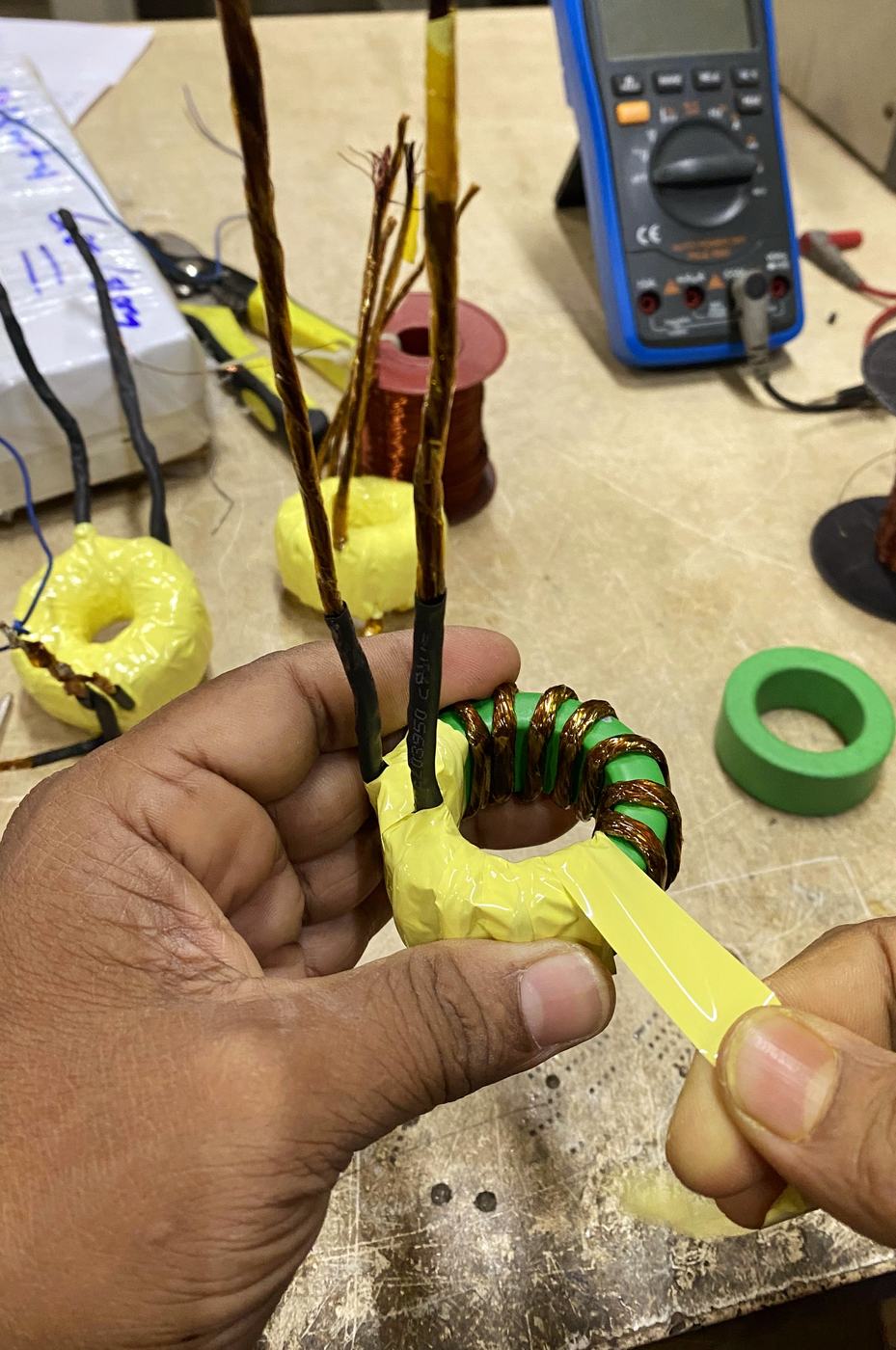 Hands wrapping yellow insulating tape around a copper winding on a green toroidal core
